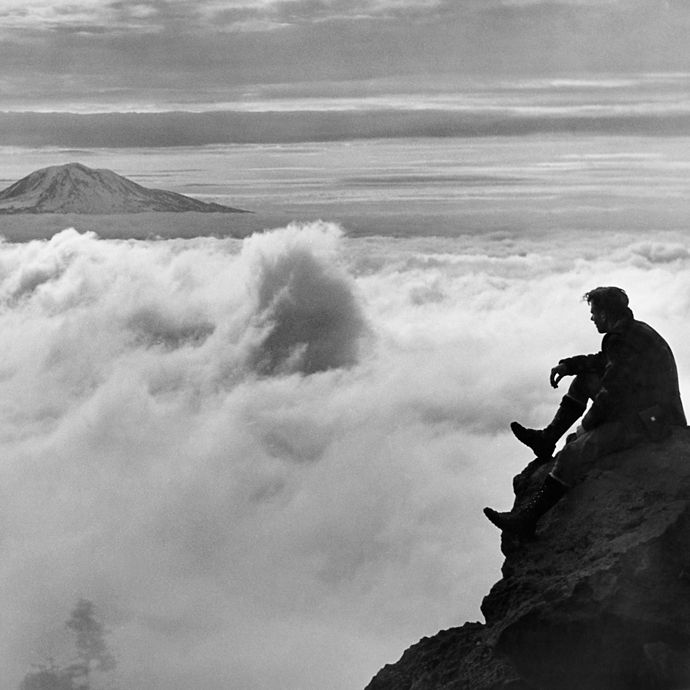 A person sitting on the edge of a rock cliff looking out over the clouds at a mountain peak in the distance.