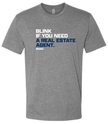 Gray short‑sleeve T‑shirt with the text “BLINK IF YOU NEED A REAL ESTATE AGENT.” printed in white and blue, with the RE/MAX logo beneath the text.