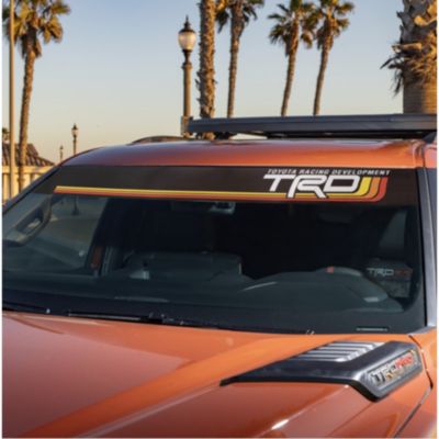 Front windshield of an orange vehicle with a black sun visor strip featuring ""TRD"" in white letters and colorful stripes.