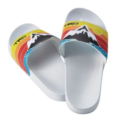 A pair of white slide sandals with colorful straps. The straps feature a design with a mountain, the sun, and rainbow-like stripes in red, orange, and yellow. The letters 'TRD' are printed in black on the left side of each strap