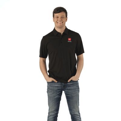Person in a black polo shirt with a small red and white logo on the chest, wearing blue jeans and standing with hands in pockets;