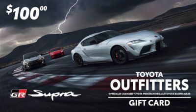 $100 Toyota Outfitters gift card with three Toyota Supra cars racing. Stormy sky background. Text reads 'Toyota Outfitters' and 'Officially licensed Toyota merchandise and Toyota racing gear.' GR Supra logo.