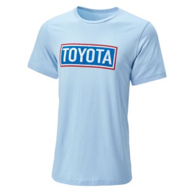 A light blue short-sleeve T-shirt with the word 'TOYOTA' printed in bold letters inside a rectangular box on the chest. The text is white with a red outline, and the box has a blue border.