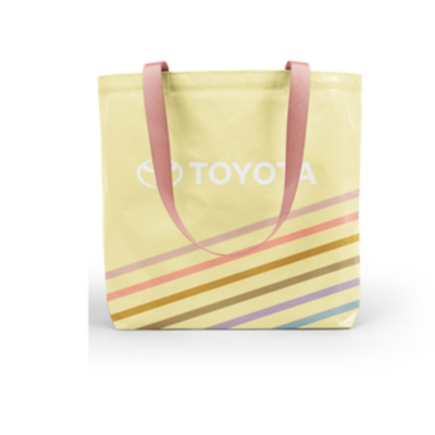 A yellow tote bag with pink handles featuring the Toyota logo in white. The bottom half of the bag has diagonal stripes in various colors including brown, yellow, orange, purple, and blue