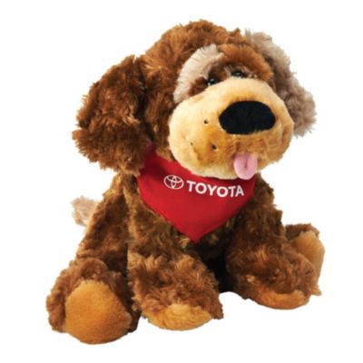 A plush toy dog with brown fur, wearing a red bandana that has the Toyota logo and text on it. The dog has floppy ears, a black nose, and its tongue sticking out.