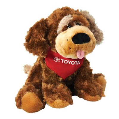 A plush toy dog with brown fur, wearing a red bandana that has the Toyota logo and text on it. The dog has floppy ears, a black nose, and its tongue sticking out.