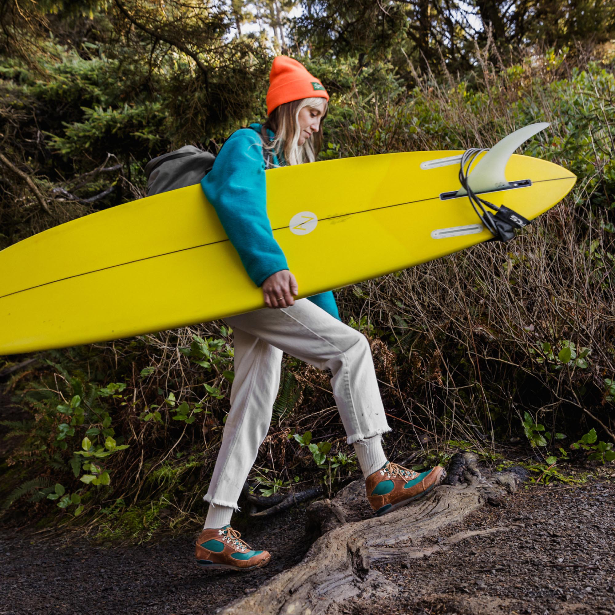 A woman walks up a trail carrying a yellow surf board with lush greenery in the background.