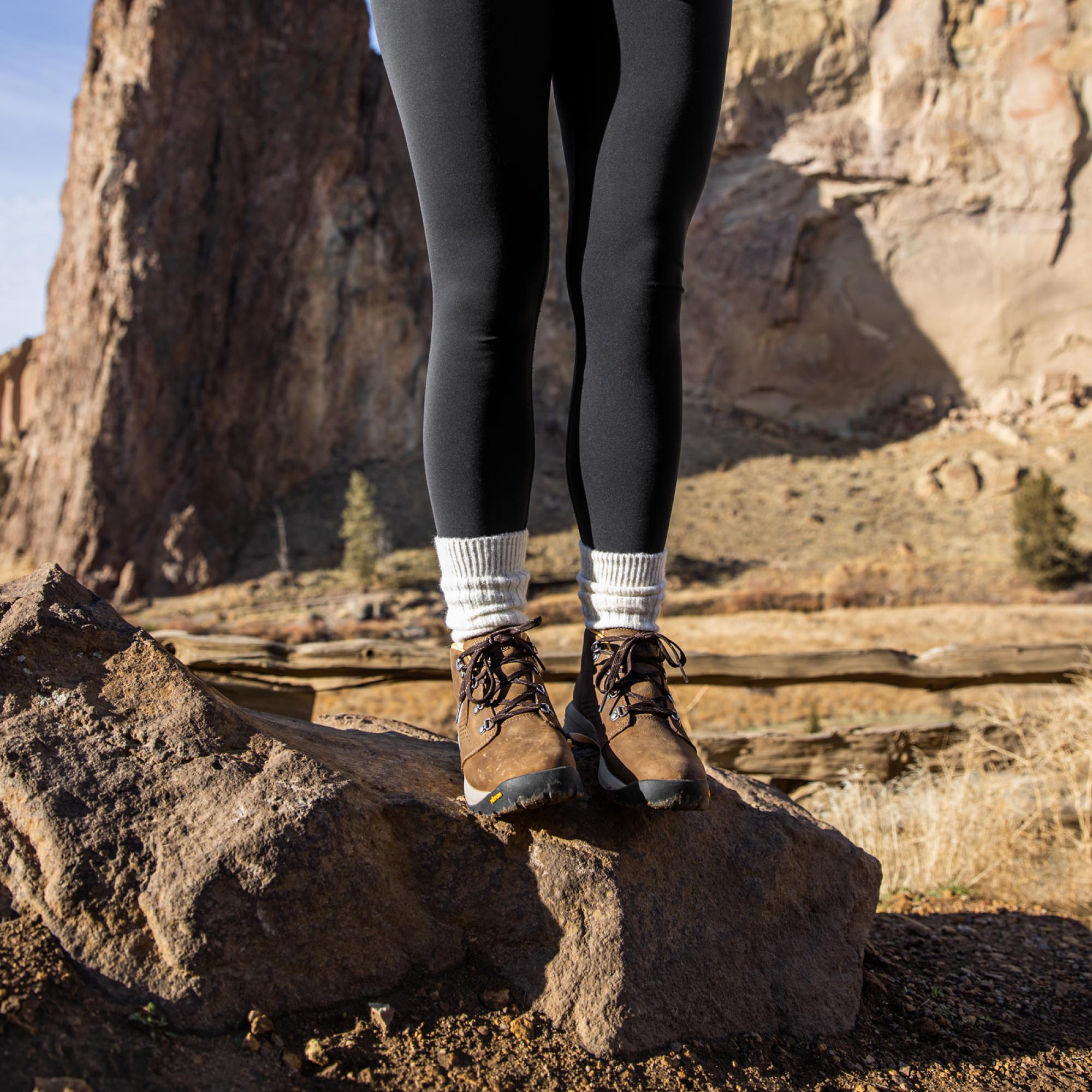 A woman stands on a rock with her hands in her yellow jacket wearing black leggings and brown leather boots with a sharp rock face in the background.