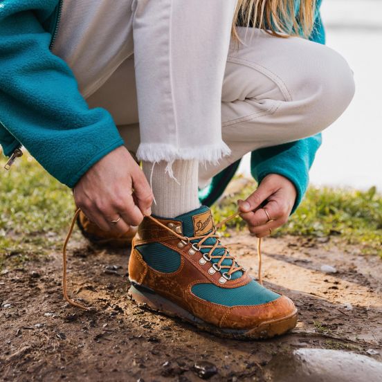 A woman bends down on a dirt trail to tie a brown leather boot with teal colored textile inlays, her pants and socks are white and her fleece top matches the teal of the boot.