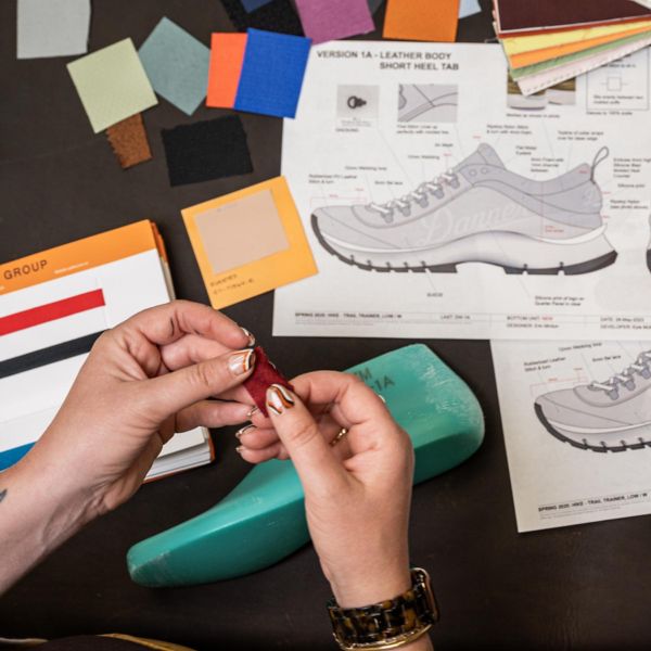 Two pairs of hands compare color swatches over a desk with a boot last, color swatches, and technical boot drawings in the background.