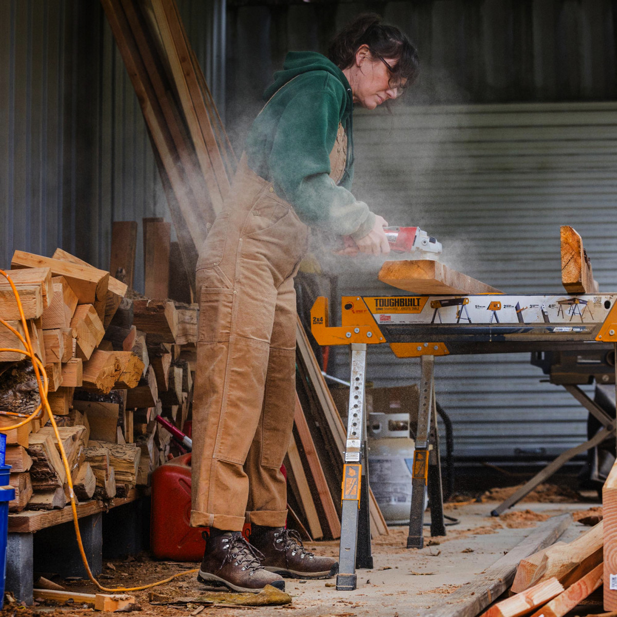 A woman dressed in overalls and brown leather boots uses a rotary sander on a rough cut wood 2x6 resting on saw horses in a covered workshop.