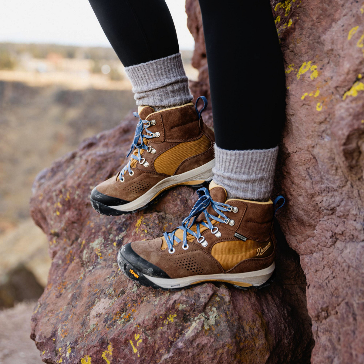 A woman pauses on a rock while hiking wearing black leggings, gray socks, and brown leather boots with yellow textile inlays and light blue laces.