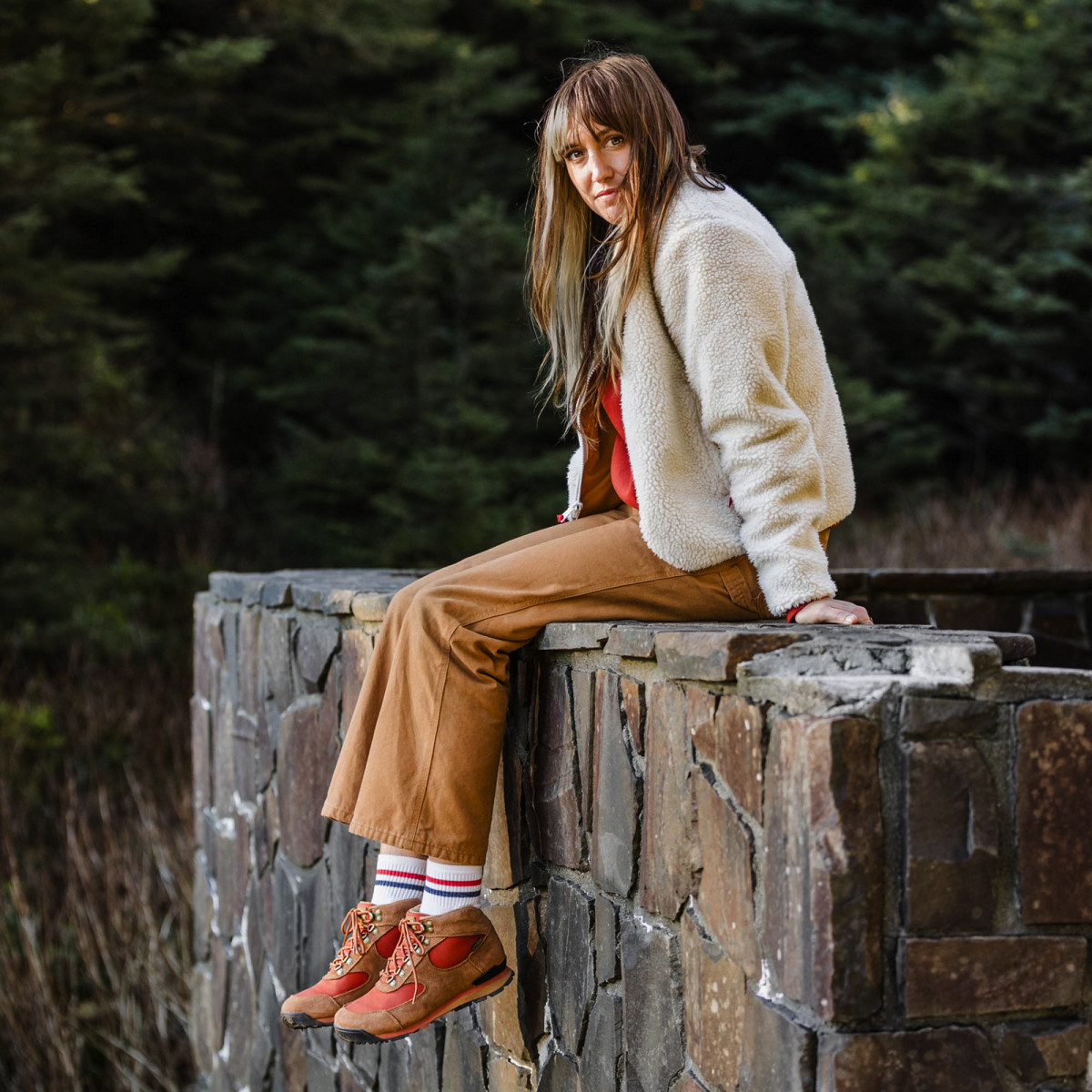 A woman sits on a rock wall, her legs dangling off the edge, wearing a white fleece top, tan pants, and brown leather boots with red textile inlays.