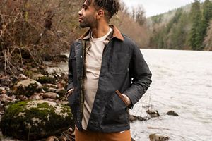 a man walks alongside a river wearing a dark waxed canvas jacket