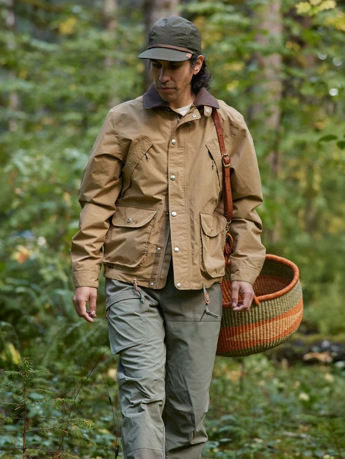 Two people walk on a well-maintained loamy trail through a lush, dark forest wearing earth-toned classically styled clothing and boots, their calm, smiling faces suggest they’re enjoying their walk and each other