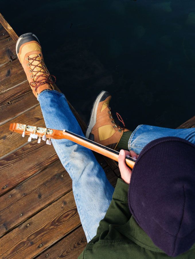 A pair of light brown leather boots with red laces rest on a pine bough with a metal mug and silverware nearby, suggesting a restful moment at camp after a hike