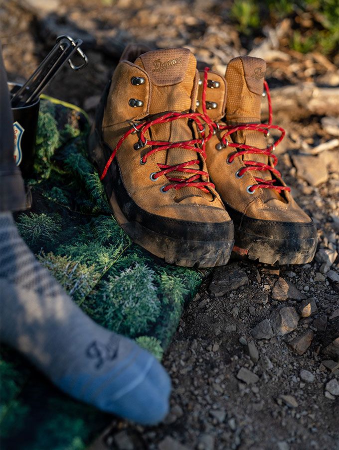 A pair of light brown leather boots with red laces rest on a pine bough with a metal mug and silverware nearby, suggesting a restful moment at camp after a hike