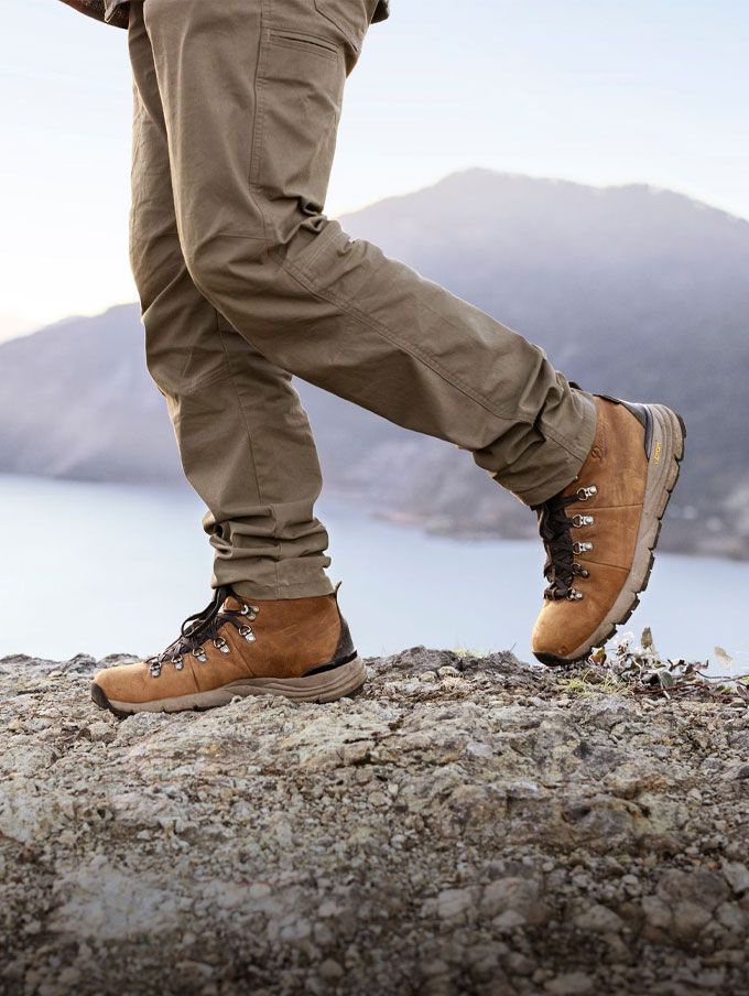 A hiker stands on a rock wearing gray boots illuminated by low evening sunlight in a dry landscape, a pause after a long day on the trail