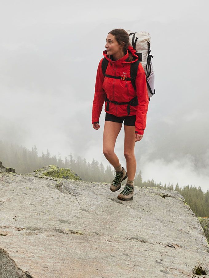 A hiker climbs rocky terrain with clouds and trees visible in the background below