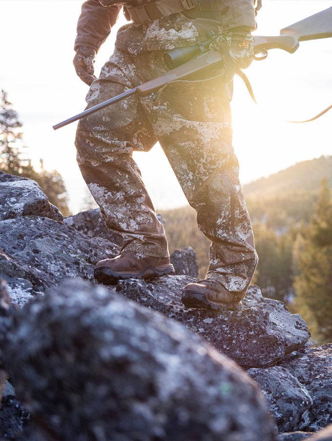 A hunter adorned in camo carries his rifle up a steep rocky hill