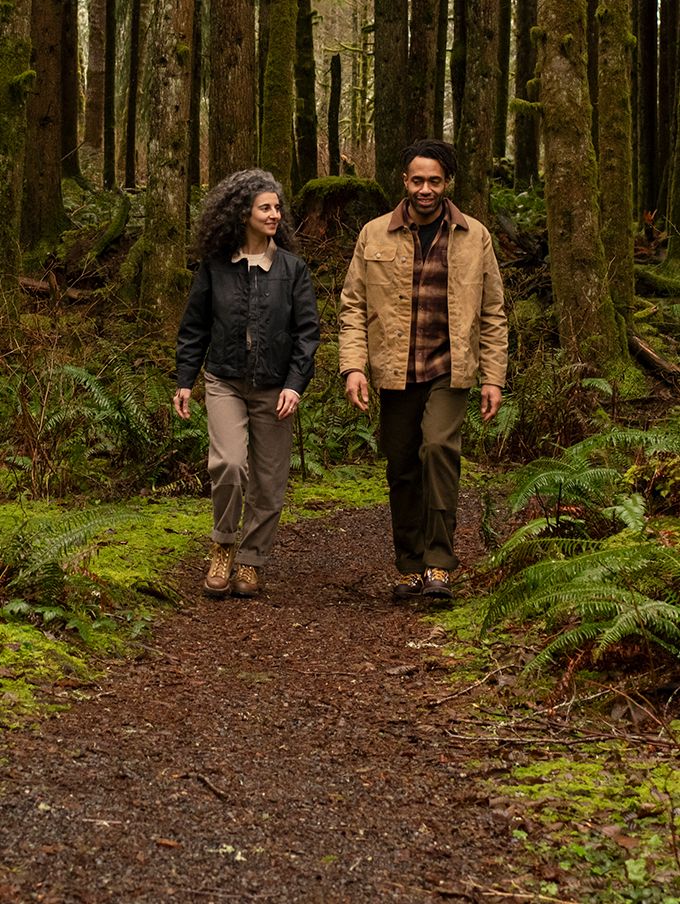 Two people walk on a well-maintained loamy trail through a lush, dark forest wearing earth-toned classically styled clothing and boots, their calm, smiling faces suggest they’re enjoying their walk and each other