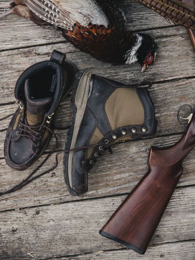 Brown leather and textile boots rest, unlaced, on a weathered wood porch next to a deceased pheasant and a rifle, the scene suggests it was a successful hunting day