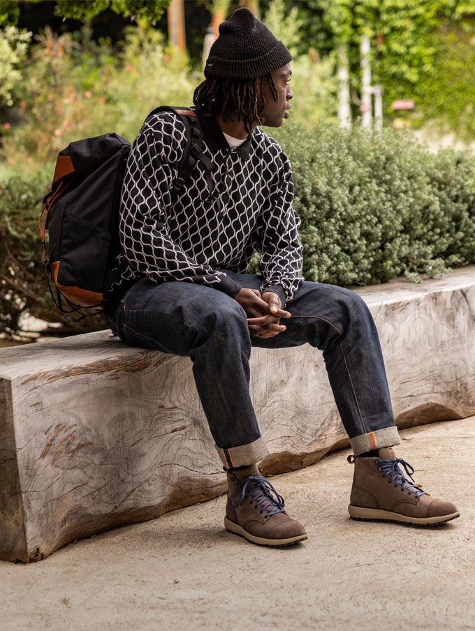 A stylishly dressed person with a backpack casually slung over one shoulder and tidily tied boots sits on a weathered wooden bench on a sunny day, the scene suggests they feel prepared for whatever their day has in store