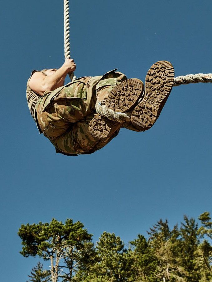 A stong-looking person suspends from a thick rope wearing camo pants, the rope is weaved around one foot showing the grippy outsole tread suggesting their boots are a key tool for their job