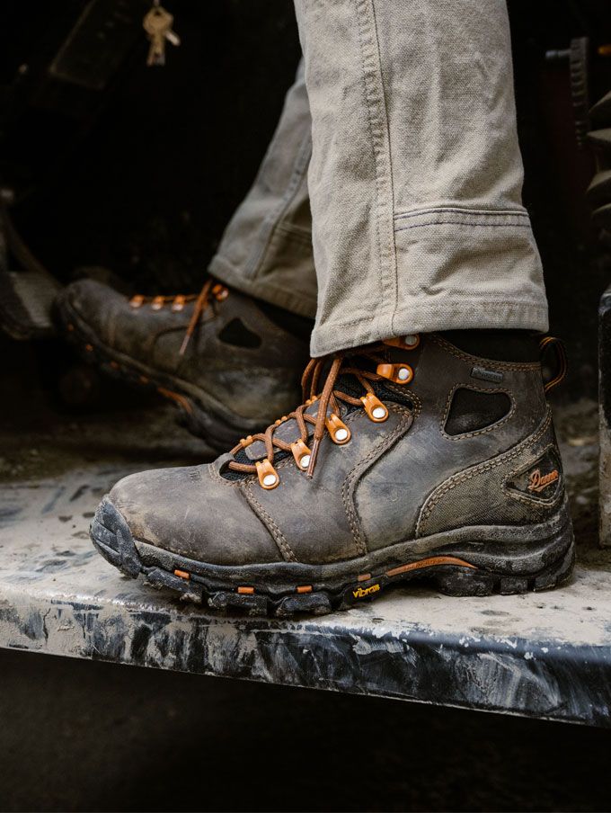 Muddy brown work boots with orange eyelets and laces rest on a red metal grate suggesting a good day of work