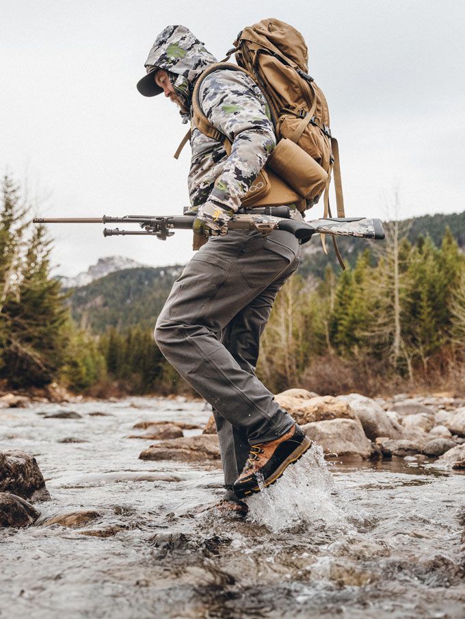 a man in a camo jacket walks through a stream while wearing brown leather hunting boots with a black rubber rand