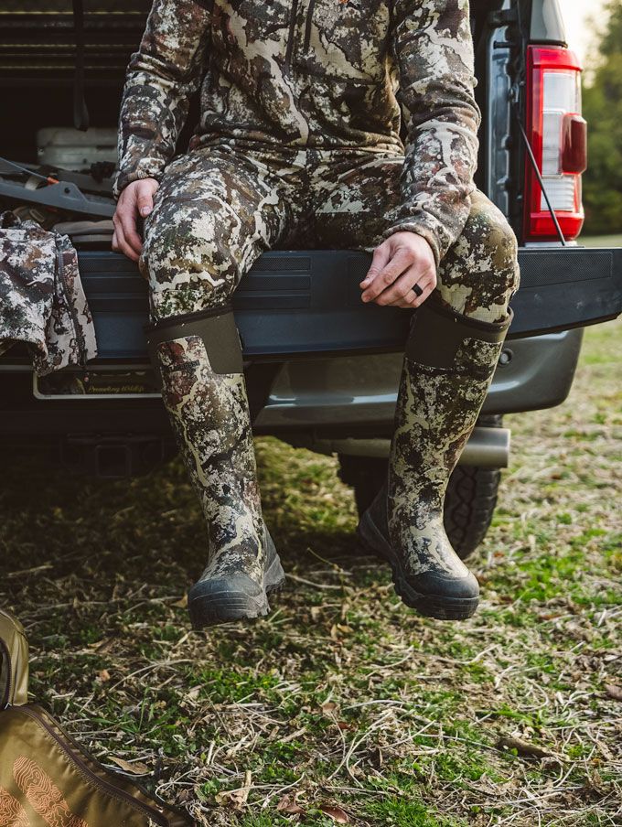 a man sits in the back of his truck adorned in camo and wearing knee high rubber boots