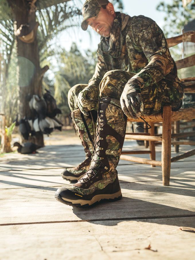 a man in camo sits on the porch of a cabin while lacing up his knee high leather and nylon snake boots