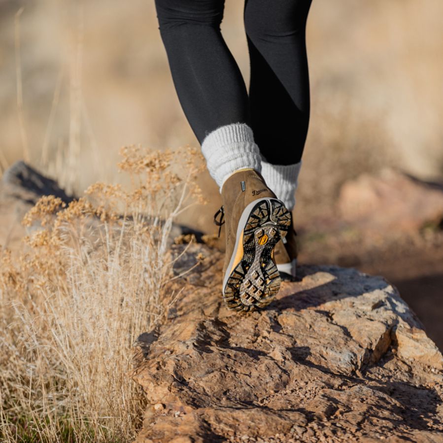 A woman walks away from the camera on a rock path wearing brown leather boots, white socks, and black leggings, the grasses are dry and tan colored in the foreground and background.