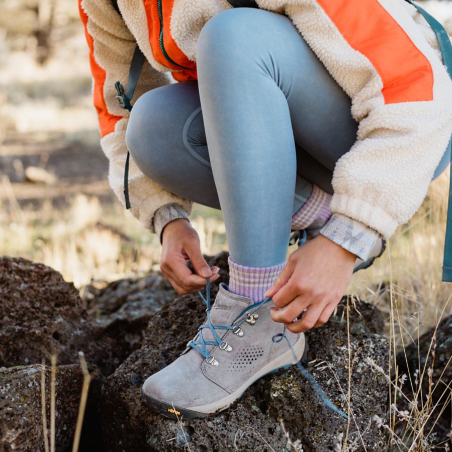 A woman pauses on a rock to tie her light gray leather boot with blue laces.