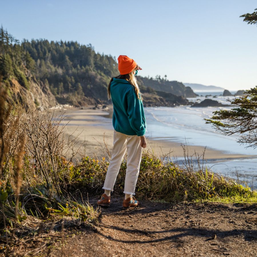 A woman pauses on a trail to gaze out at the ocean on a sunny day with an evergreen covered peninsula in the background.