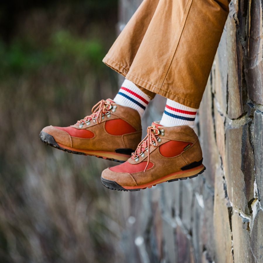 The lower half of a woman’s legs dangle off a rock wall wearing brown pants, white socks with stripes, and brown leather boots with red textile inlays.