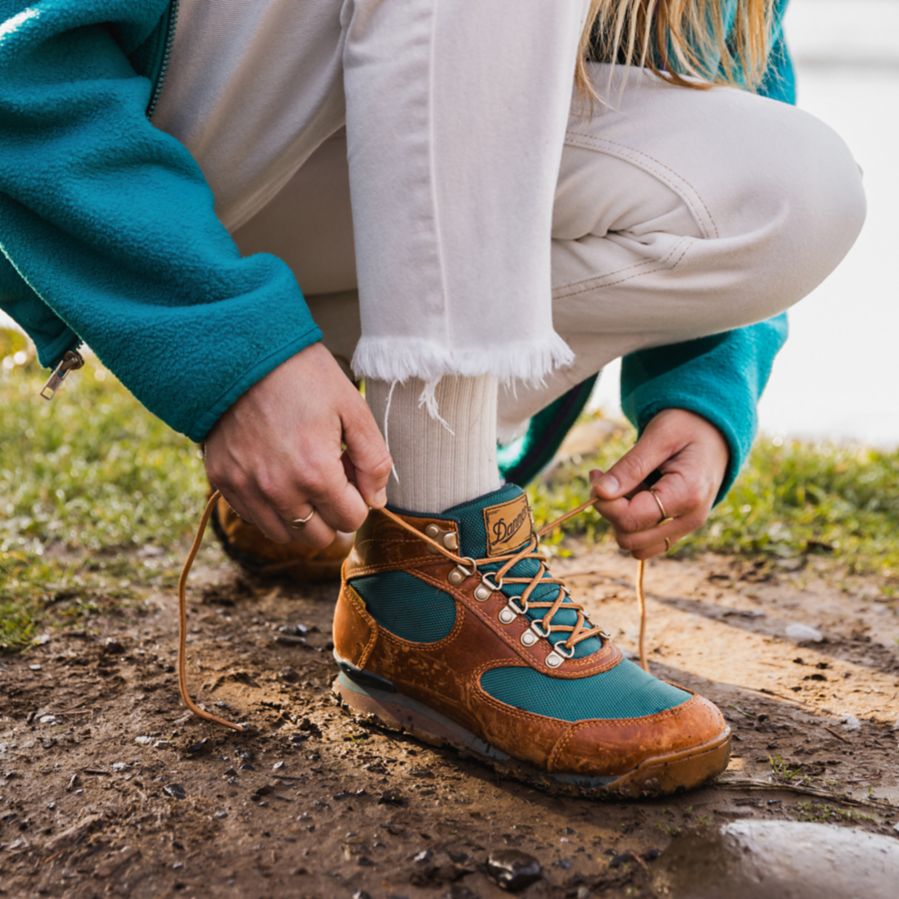 A woman bends down on a dirt trail to tie a brown leather boot with teal colored textile inlays, her pants and socks are white and her fleece top matches the teal of the boot.