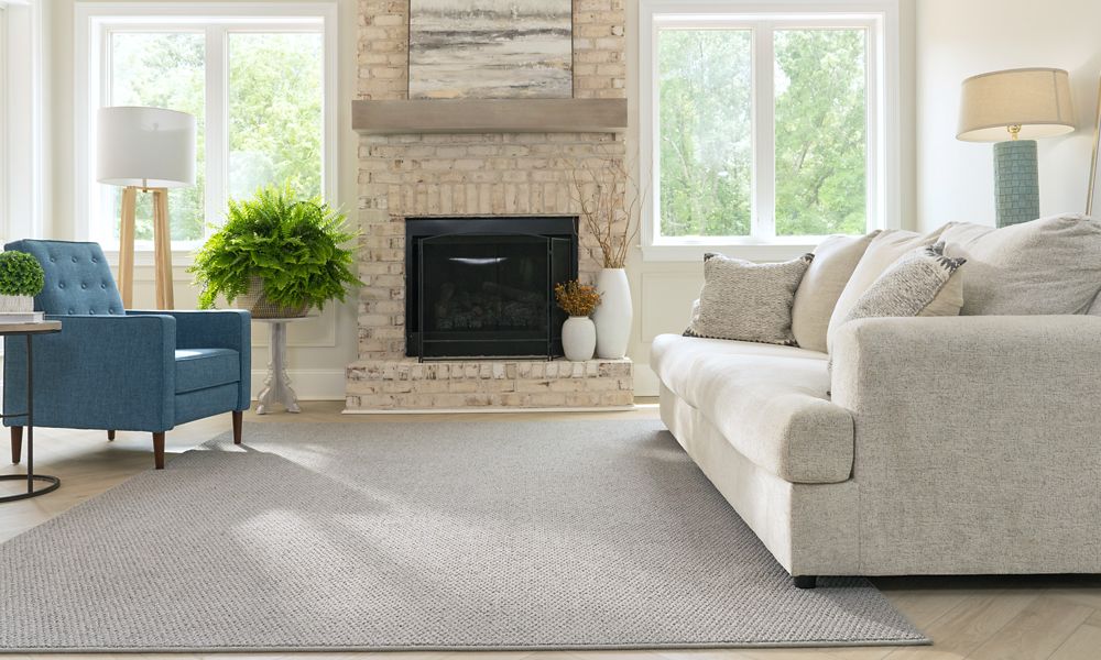 A sunny living room with a grey couch, blue chair, fireplace, and a grey carpet