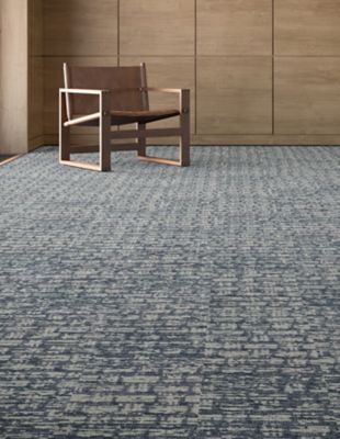 A contemporary armchair, featuring a tan leather seat and wooden frame, is positioned on a Crafted Expression carpet tile in color 565 Rabbitbrush, complemented by the room's light wood paneling.