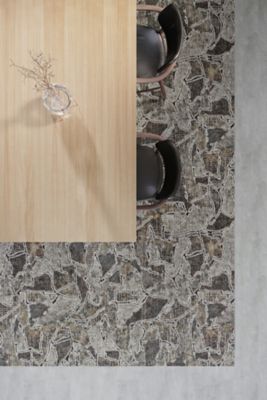 A top-down image of a rectangular dining table with a light woodgrain finish. Two black chairs with wooden arms are arranged around the table, which is set upon a Innate Spirit area rug. The rug is placed on Bolder 2.5|5.0 LVT in 929 Celestial.