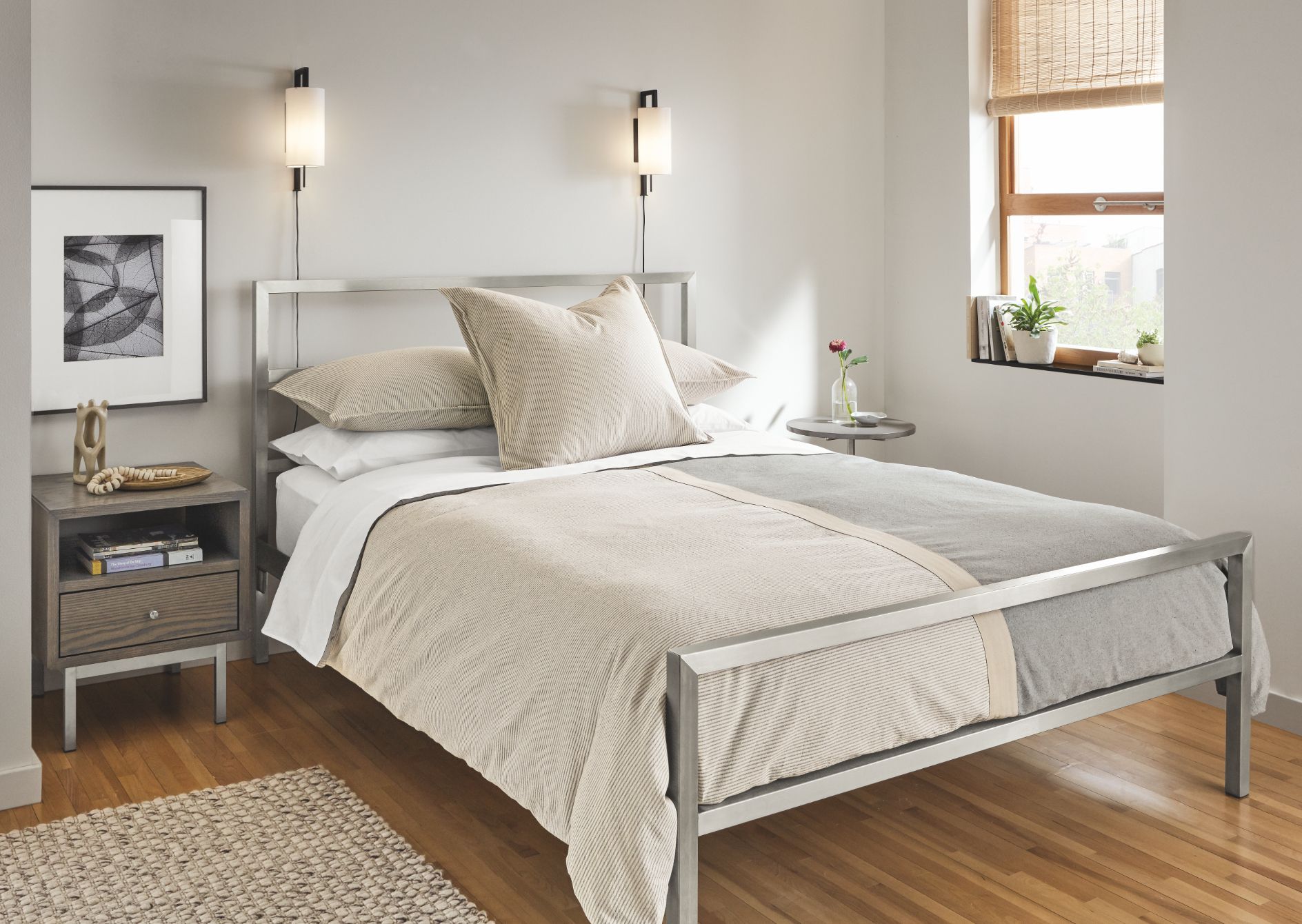  A small bedroom with a bed, a nightstand, and a small table with a plant on it. The furniture is all made of light wood and the walls are white.